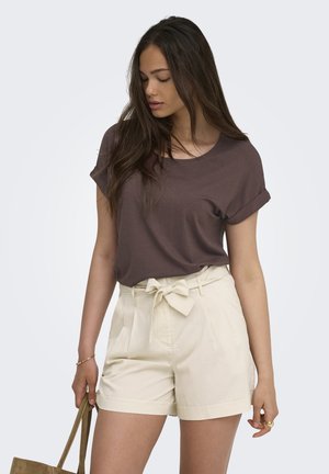 Young woman wearing a brown short-sleeve top and high-waisted beige shorts with a bow belt, holding a brown handbag and looking down.