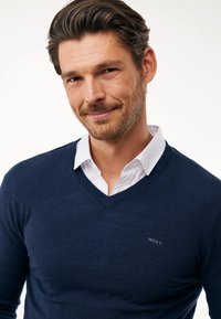 Marinblå v-ringad tröja i stickad tyg, med en slim fit och en subtil logotyp på bröstet, lagd över en vit skjorta med krage.