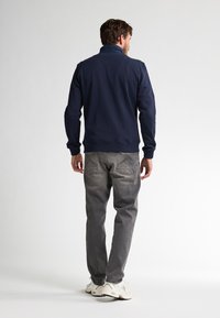 Felpa zip-up blu navy con colletto alto, abbinata a pantaloni in denim grigi e sneakers bianche, vista da dietro. Texture del tessuto liscia.