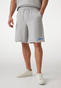 Shorts in cotone grigio con vita con coulisse, orlo grezzo e logo blu "GUESS". Indossati con sneakers bianche su uno sfondo neutro.