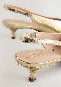 Tacchi slingback dorati con una finitura metallica screpolata, tacco basso e una soletta rosa con il marchio "NEXT" in rilievo.