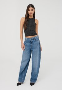 Młoda kobieta z długimi brązowymi włosami, ubrana w bezrękawnikowy, czarny top z ćwiekami, luźne niebieskie dżinsy i czarne, spiczaste buty, stojąca na białym tle.