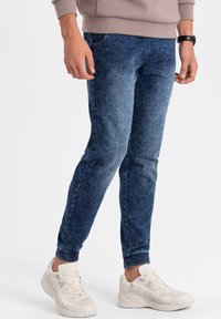 Joggers en denim bleu foncé avec un délavage, poignets élastiques et coupe régulière, assortis à des chaussures de sport claires.
