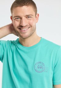 T-shirt en coton turquoise à manches courtes, avec un col rond et un imprimé de logo violet incluant un design de crabes.