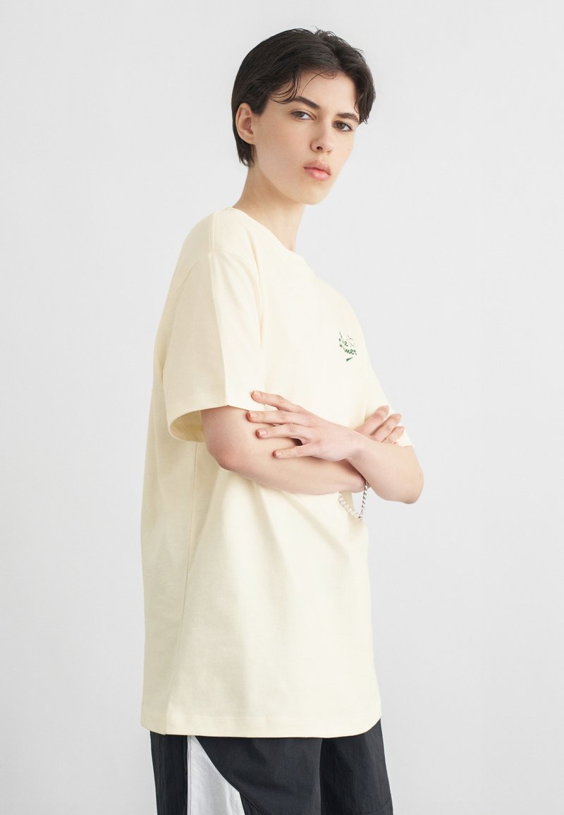 Άτομο με κοντά σκούρα μαλλιά, φορώντας υπερμεγέθη κρεμ μπλούζα και μαύρο παντελόνι, στέκεται με σταυρωμένα τα χέρια μπροστά σε απλό γκρι φόντο.