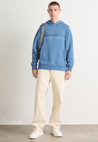 Felpa in cotone blu con logo "HUGO" ricamato in colore contrastante, tasca a marsupio frontale, abbinata a pantaloni larghi color panna e sneakers bianche.