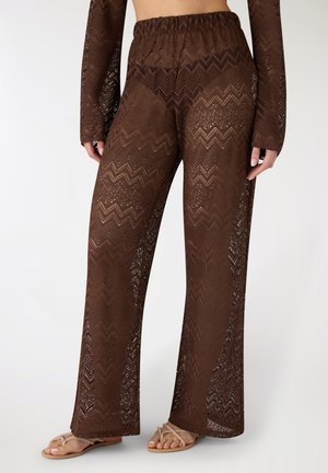 Pantaloni ampi color marrone con motivo a zigzag trasparente, vita elastica, indossati con top a maniche lunghe abbinato e sandali beige.