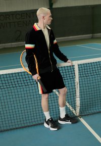 Satiinmustast spordikomplektist koosnevad mitmevärvilised triibud, mustad lühikesed püksid ja Nike jalatsid. Hoides tennisereketit võrgu lähedal.
