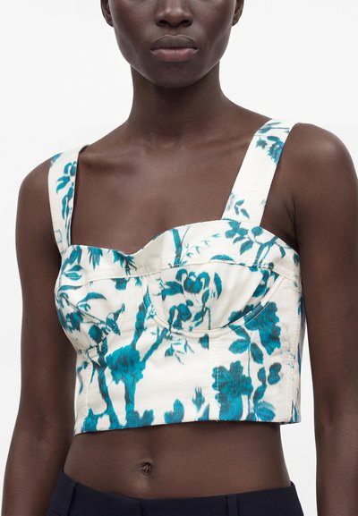 Personne à la peau foncée portant un haut court blanc avec de larges bretelles et un imprimé floral bleu, associé à un pantalon sombre, vue du cou à la taille.