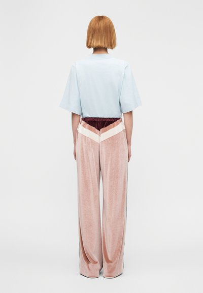 Άτομο με κοντά κόκκινα μαλλιά που φοράει ανοιχτό μπλε φαρδιά κοντομάνικη μπλούζα και άνετο ροζ βελούδινο παντελόνι με λευκό και μπορντό σχέδιο σε σχήμα έψιλον.