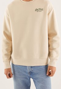 Felpa beige con collo a giro, caratterizzata da una scritta ricamata verde "Club House MIAMI BEACH, FLORIDA" e una texture morbida. Abbinata a jeans blu.