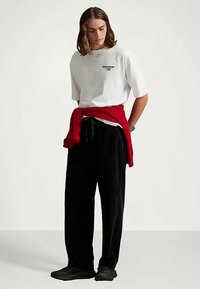 Ung man med axellångt hår iförd en vit t-shirt med logotyp, svarta byxor, svarta skor och en röd tröja knuten omkring midjan, stående.