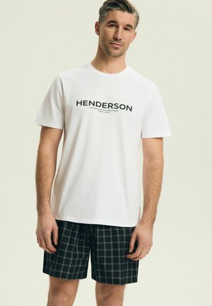 Mężczyzna w białej koszulce z logo Henderson i czarnych szortach w kratę stojący na tle jasnego, jednolitego tła.