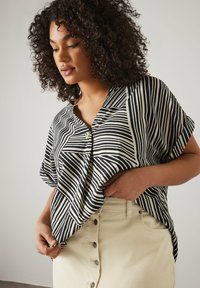Blusa a maniche corte a righe bianche e nere con dettaglio dei bottoni, abbinata a una gonna beige chiaro con una fila di bottoni.