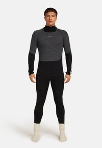 Un uomo sta in piedi di fronte, indossando una maglia a maniche lunghe a due tonalità, grigia e nera, leggings neri e calzini di colore crema.