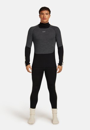 Un homme se tient face à l'avant, portant un haut ajusté à manches longues gris et noir à deux tons, des leggings noirs, et des chaussettes de couleur crème.