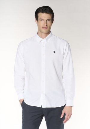 Uomo con capelli scuri che indossa una camicia bianca con bottoni e un piccolo logo ricamato sul petto e pantaloni scuri, in piedi con le mani nelle tasche.