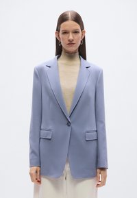 Ανοιχτό μπλε σακάκι με πέτο γιακά, κλείσιμο με ένα κουμπί και δύο μπροστινές τσέπες. Φοριέται πάνω από ένα μπεζ ζιβάγκο, με λείο υφασμάτινο υλικό.