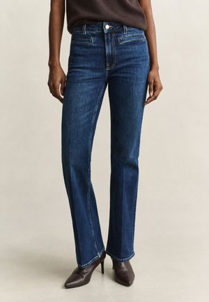 Femme portant un jean bleu foncé droit et des bottes marron à talons, debout avec les mains détendues le long du corps.