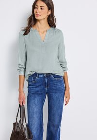 Blouse vert clair avec un col en V, manches froncées, associée à un jean large bleu. Le mannequin tient un sac en filet marron.