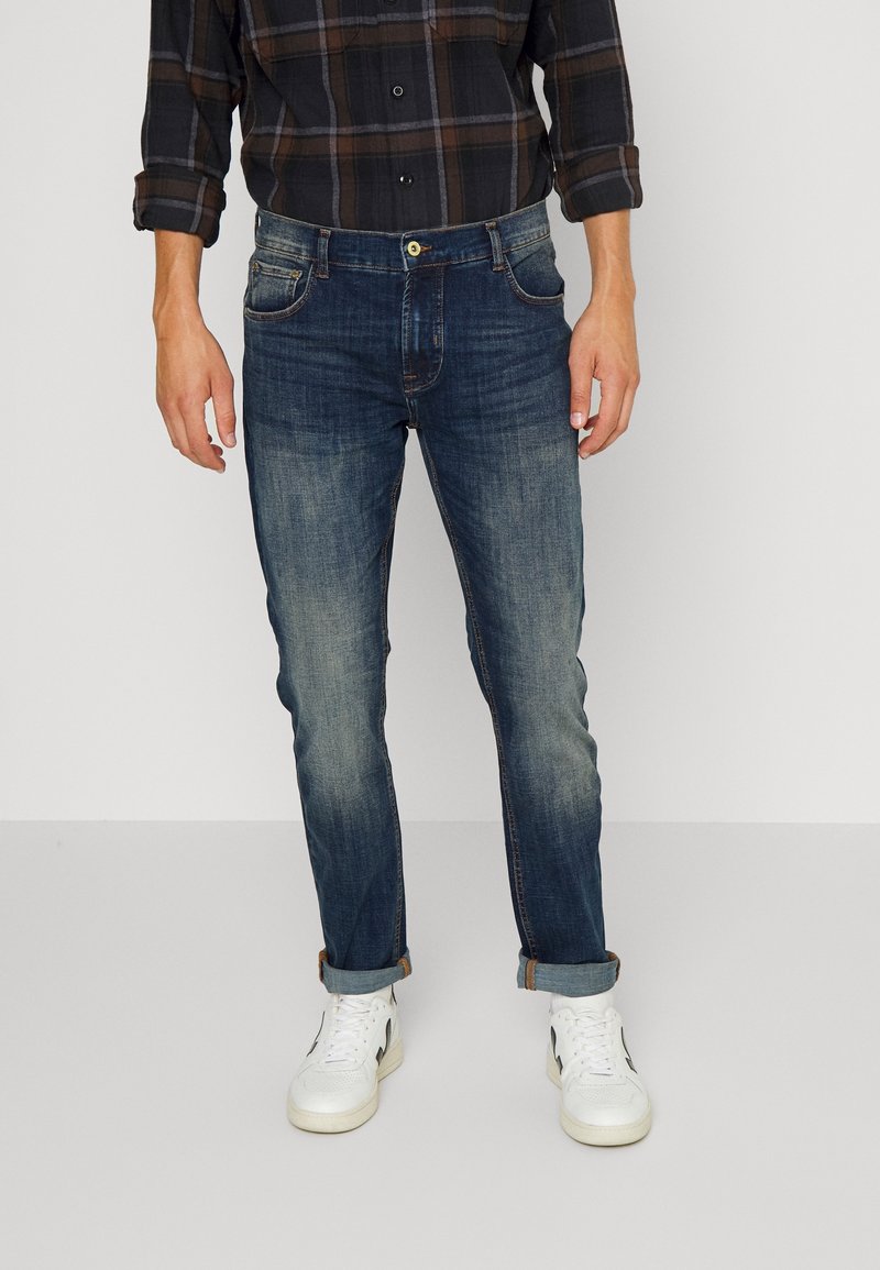 Homme portant un jean en denim bleu foncé retroussé, des baskets blanches et une chemise à carreaux foncée avec les manches retroussées, debout devant un fond uni.
