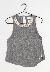 Top sans manches gris avec une finition texturée, arborant un col en perles rappelant des perles de culture. Ourlet arrondi avec de légentes fentes sur les côtés, suspendu à un cintre noir.