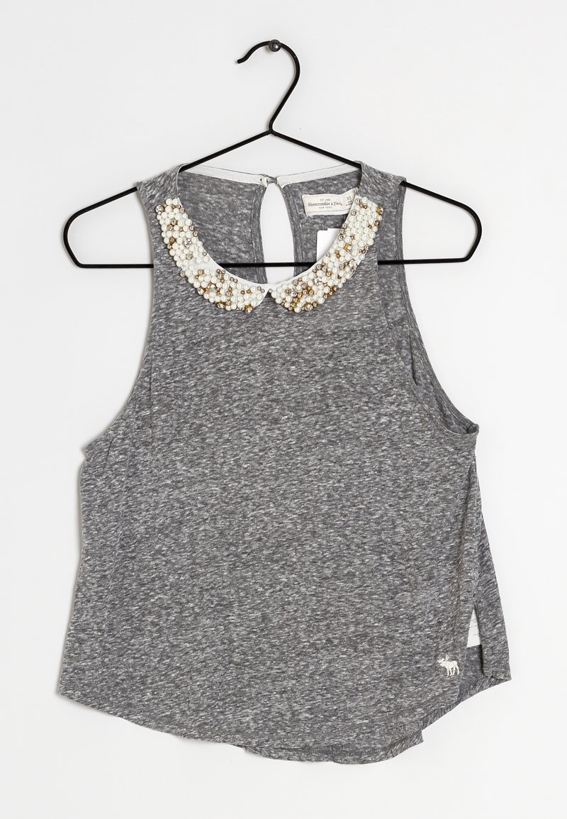 Top sans manches gris avec une finition texturée, arborant un col en perles rappelant des perles de culture. Ourlet arrondi avec de légentes fentes sur les côtés, suspendu à un cintre noir.
