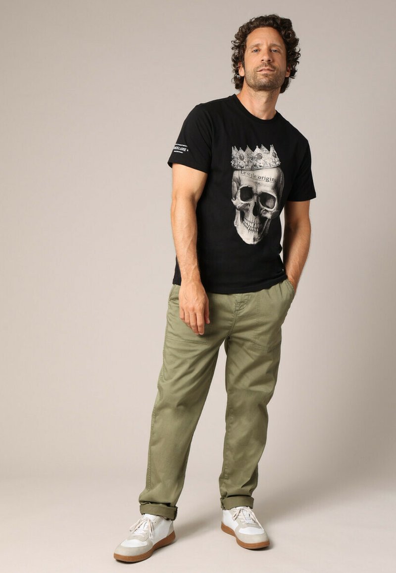 T-shirt en coton noir avec un graphique de crâne portant une couronne, associé à un pantalon vert olive clair et des baskets blanches avec des semelles marron.