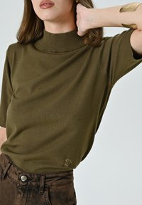 Donna che indossa un pullover a collo alto a maniche corte verde oliva e pantaloni marroni a vita alta, con un braccio alzato dietro la testa.