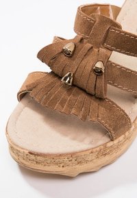 Sandale compensée en daim marron avec pompons à franges et embouts métalliques dorés sur semelle en liège, avec doubles brides et coutures visibles.