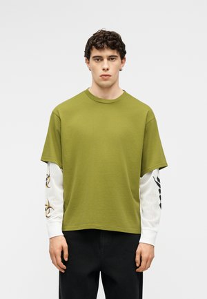 Hombre joven con cabello rizado corto que lleva una camiseta verde de manga corta sobre una camisa blanca de manga larga con símbolos negros y dorados.