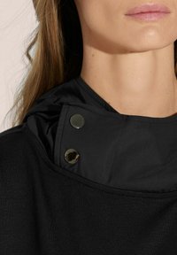 Zwarte kledingstuk met een hoge kraag, voorzien van twee metalen drukknoopjes en textiel met een textuur bij de hals. Over het geheel een gladde, zachte textuur.