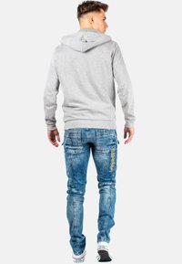 Felpa grigia con cappuccio e tasca a marsupio, abbinata a jeans blu scuro strappati con più tasche e dettagli in cucitura gialla.