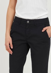 Person's torso wearing a white ribbed top and black pants with a button, belt loops, and hands resting at sides.