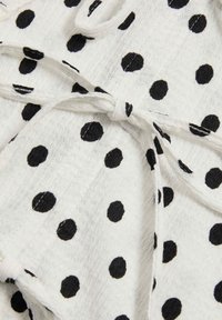 Witte stof met zwarte polka dots, met een gestructureerd patroon. Strikbanden zijn aanwezig, wat zorgt voor een betere pasvorm van het kledingstuk.