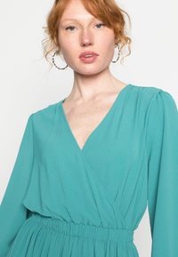 Vestido envolvente azul-petróleo com decote em V e mangas longas, apresentando uma cintura elástica e um tecido macio e fluido. Design texturizado e sem forro.