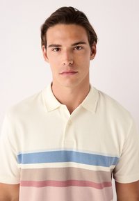 Polo color crema con strisce orizzontali in azzurro chiaro, bianco, rosa tenue e beige. Dotata di colletto e patta con tre bottoni.