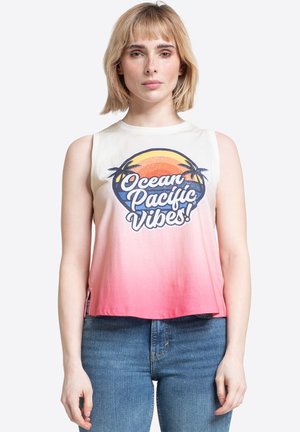 Kobieta o blond włosach, ubrana w bezrękawnik ombre z napisem „Ocean Pacific Vibes!” i grafiką palmy, w połączeniu z niebieskimi dżinsami.