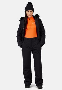 Giacca puffer nera calda con cappuccio bordato di pelliccia, maglietta arancione a maniche lunghe e pantaloni da sci neri con vestibilità comoda e dettagli con zip.