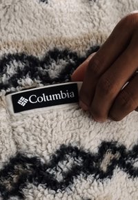 Main tenant une étiquette de marque Columbia sur un tissu polaire à motifs crème et noir.