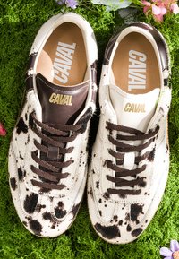 Paire de baskets Caval noires et blanches tachetées avec des lacets marron posée sur de la mousse verte avec des fleurs roses et violettes à proximité.