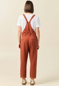 Rostfärgade hängselbyxor med vida ben, justerbara axelremmar, bakfickor och uppvikta fållar, bärs över en vit t-shirt. Slät tygtextur.