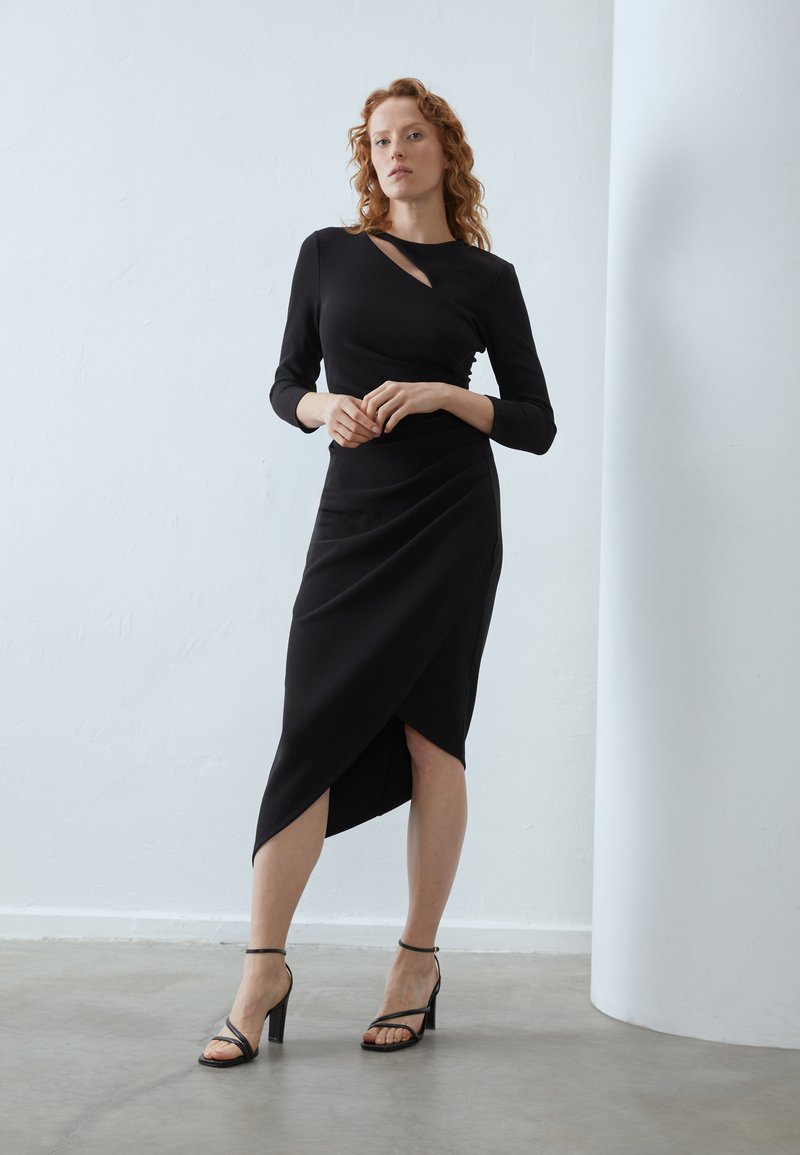 Schwarzes Wickelkleid mit langen Ärmeln, das einen diagonalen Ausschnitt am Hals und einen hohen Schlitz aufweist, kombiniert mit schwarzen Riemchen-Heels.