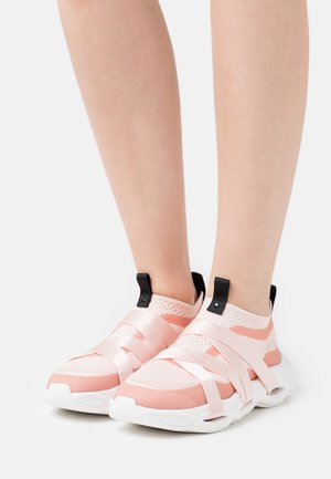 Jambes portant des chaussures de sport légères roses pâles à enfiler avec des semelles blanches et de larges brides en satin croisant le dessus, ainsi que des languettes noires à l'arrière.
