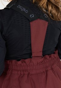 Salopette bordeaux con spalline nere regolabili, caratterizzate da un logo a zigzag e un tessuto testurizzato nella parte superiore. Dettagli della vita arricciata visibili.