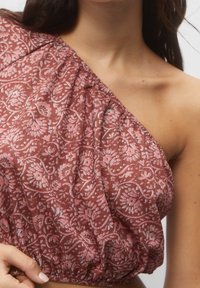 Haut court asymétrique en tissu texturé couleur rouille avec motif floral en rose clair, présentant un détail froncé à l'ourlet et une finition douce.