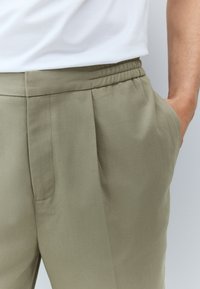Light olive green trousers with side pockets, a relaxed fit, and a pleated front design. Soft fabric with a smooth texture.