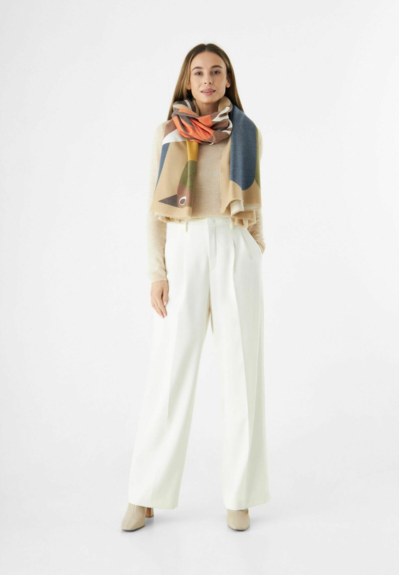 Écharpe à motifs multicolores drapée sur un pull léger ; associée à un pantalon large blanc et des bottillons neutres. Texture lisse, design moderne.