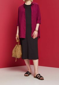 Femme portant une robe midi noire et une chemise magenta déboutonnée, tenant un sac en osier et portant des sandales plates noires à brides.