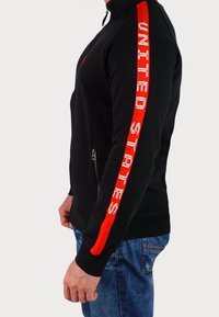 Felpa nera con zip e collo alto; presenta una striscia rossa e il testo "UNITED STATES" lungo la manica destra; tessuto testurizzato.